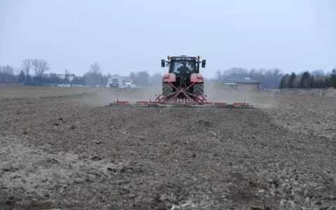 Czerwony traktor z agregatem uprawowym przygotowuje pole pod zasiew. Dzierżawa ziemi w pełni.