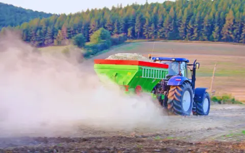 Traktor rozsiewa wapno do ziemi, tworząc chmurę pyłu na polu. W tle las i wzgórza.