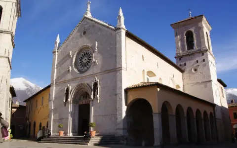 A grand church with a rose window and bell tower stands in a town square, with snow-capped mountains in the distance. This is Nursia, a historic Italian town.