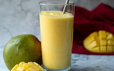 Orzeźwiający koktajl z mango w szklance, obok świeże mango i kawałki owocu. Idealny na upalne dni.