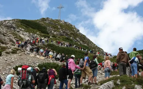 Tłum turystów wspina się na Giewont, podziwiając widok z Kasprowego Wierchu. Na szczycie widoczny krzyż.