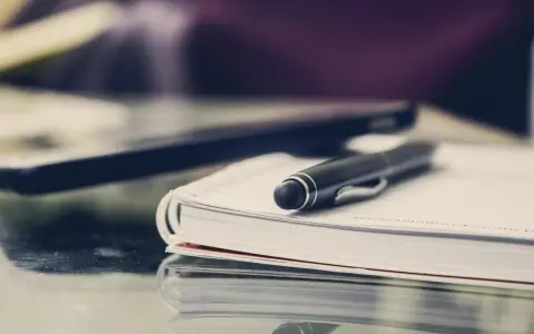 Czarny długopis spoczywa na otwartym notatniku. Obok leży laptop, gotowy do pracy. FGŚP.