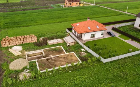 Działka 1000m2 wymiary: rozpoczęta budowa domu, gotowy fundament, sterta cegieł, drewno, żwir, obok stoi dom z pomarańczowym dachem i samochód.