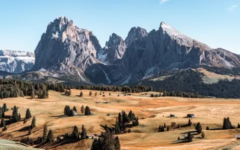 Un paesaggio mozzafiato delle Dolomiti più belle, con cime imponenti, prati dorati e boschi verdi. Piccole baite sparse creano un'atmosfera idilliaca.
