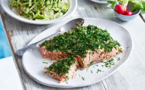 En saftig bit confiterad lax, generöst toppad med färska örter, serveras på en vit tallrik. Bredvid syns en fräsch sallad och några röda rädisor.