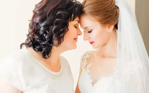 Matka i córka, obie elegancko na ślub córki, dzielą wzruszający moment. Panna młoda w białej sukni, mama w ozdobnej bluzce.
