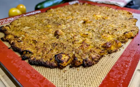 Une galette complète au four, dorée et croustillante, sur un tapis de cuisson en silicone. Des tomates jaunes et une courgette sont floues en arrière-plan.