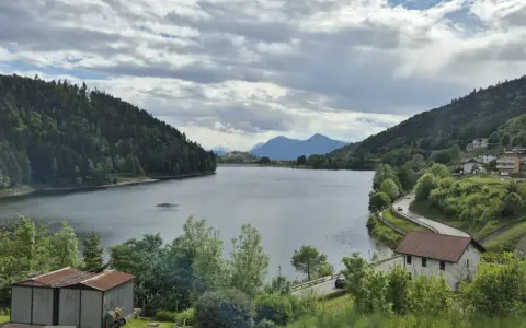 Panoramica del lago di Stramentizzo circondato da montagne verdi e un villaggio pittoresco. Nuvole minacciose nel cielo.