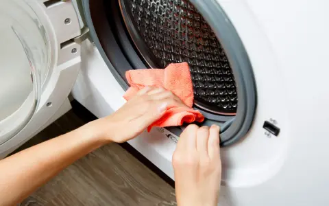Hands cleaning the rubber seal of a washing machine with an orange cloth. This is part of how to clean washing machine.
