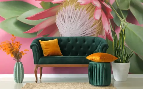 Sofá verde esmeralda con cojines naranjas, junto a un jarrón de cactus y plantas. El fondo es un mural con grandes estampados florales y hojas verdes sobre rosa vibrante.