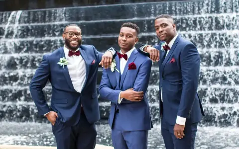 Three groomsmen in stylish navy blue suit combinations, featuring burgundy bow ties and pocket squares, pose in front of a waterfall.