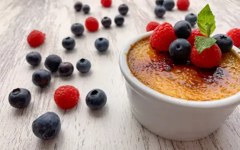 Creme brulee mit karamellisierter Kruste, garniert mit frischen Himbeeren und Blaubeeren, umgeben von weiteren Beeren auf hellem Holz.