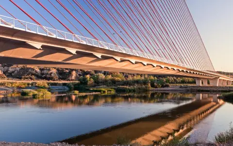El puente de Castilla-La Mancha se refleja en el río, con sus cables rojos y el cielo azul.