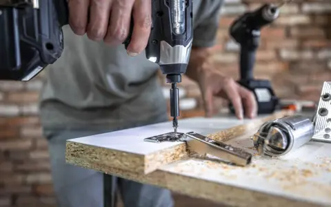 Un hombre usa uno de los tipos de taladros para atornillar una bisagra a un tablero de madera. Otro taladro y herramientas están en la mesa.