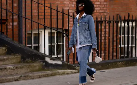 Frau in Jeans-Patchwork-Hose und Jeansjacke, ein Trend für den Jeans-Trend 2026. Weiße Sonnenbrille, weiße Sandalen.