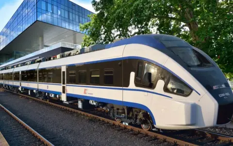 Nowoczesny, biało-niebieski pociąg Intercity ED161-002, najszybszy pociąg w Polsce, stoi na torach obok futurystycznego budynku.