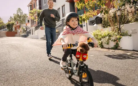 Un bambino con un mantello da supereroe prova ad andare in bicicletta, mentre suo padre corre dietro per aiutarlo. Mio figlio non riesce a pedalare, ma si diverte un mondo!