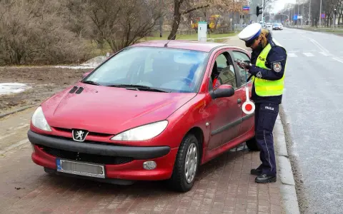 Policjantka w kamizelce odblaskowej i czapce spisuje kierowcę przy czerwonym samochodzie.