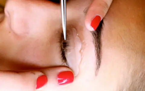 Application d'un produit pour recourber les cils. Une main aux ongles rouges tient délicatement la paupière pendant que l'outil travaille.