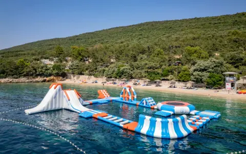 Wodne atrakcje na krystalicznie czystej wodzie, idealne na wakacje w **camping Slatina**. W tle zielone wzgórza i plaża.