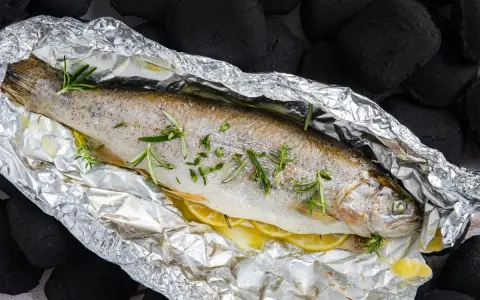 Eine ganze Forelle vom Grill in Alufolie, mit Zitronenscheiben und Rosmarin verfeinert, bereit für den Genuss.