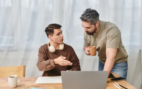 Ojciec rozmawia z synem, pokazując mu, jak zmotywować dziecko do nauki. Na stole laptop, zeszyt i kubek kawy.