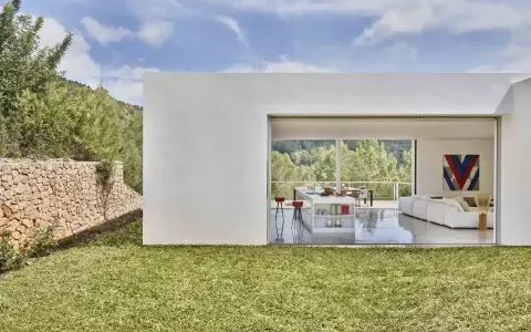 Exterior de casa ibicenca con césped verde y muro de piedra. Interior moderno con cocina y salón abiertos al paisaje.