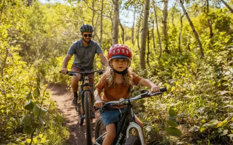 En flicka och en man cyklar på en stig i en lummig skog. Solen lyser genom träden och skapar ett vackert ljusspel.