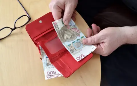 Ręce wkładają banknot 500 zł do czerwonego portfela. Obok okulary. Może to być pomocne przy sprawdzaniu 14 emerytury tabeli netto.