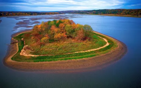 Jesienna wyspa na zalewie Brody, porośnięta kolorowymi drzewami i trawami, otoczona błękitną wodą.