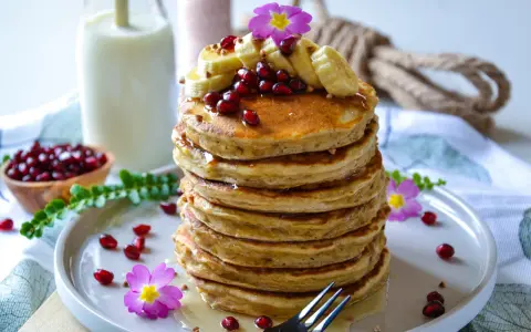 Pile de pancakes banane, garnie de fruits rouges, de rondelles de banane et de fleurs roses. Un délice pour le petit-déjeuner.