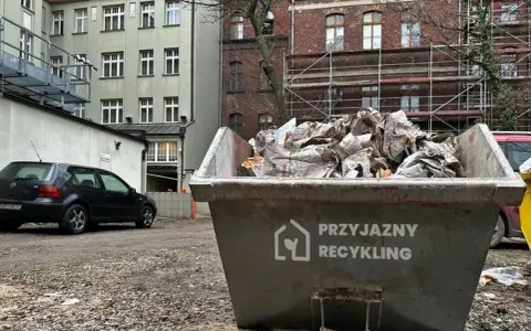 Kontener na śmieci z napisem "Przyjazny Recykling" wypełniony papierem. W tle budynki i czarny samochód. Data: 17 09 04.