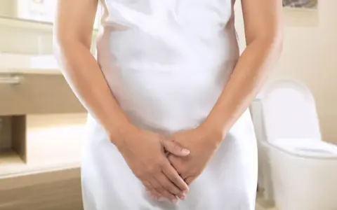 Kobieta w białej sukience w łazience, z dłońmi skrzyżowanymi na brzuchu. W tle toaleta. Szuka domowych sposobów na nietrzymanie moczu.