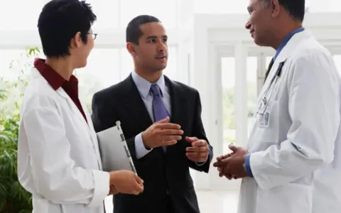 Un uomo in giacca e cravatta parla con due medici in camice bianco. Forse discutono di un nuovo farmaco e del suo informatore scientifico stipendio.