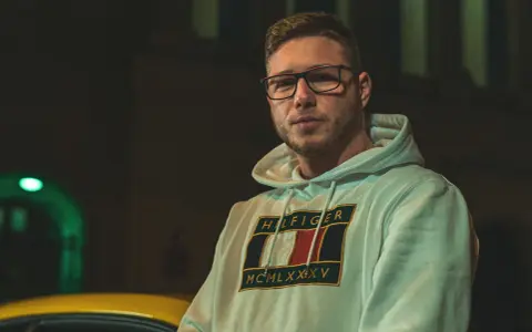 Kubańczyk cały ja utwory. Młody mężczyzna w okularach i bluzie z kapturem Tommy Hilfiger, nocą, obok żółtego samochodu.