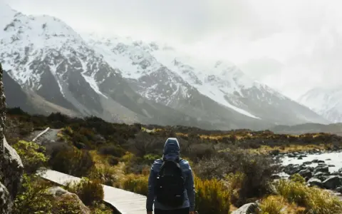 Une personne avec un sac à dos fait une randonnée sous la pluie, face à des montagnes enneigées. Le chemin en bois serpente à travers une végétation basse.