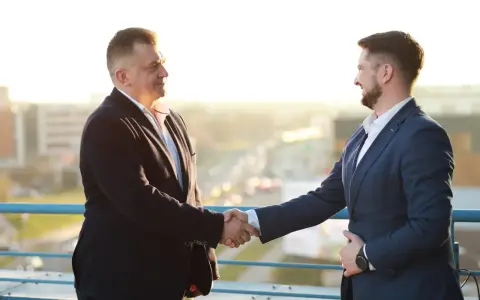 Dwóch mężczyzn w garniturach ściska sobie dłonie na tle miasta.