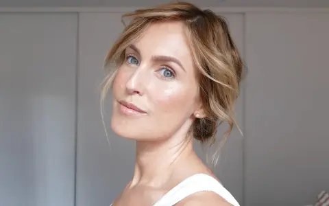 En kvinna med blont hår och blå ögon har ett naturligt glow i ansiktet. Hon bär en vit topp och en liten diamantörhänge.