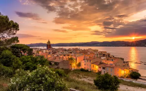 Sonnenuntergang über St. Tropez. Die Sonne versinkt im Meer, beleuchtet die Stadt und den Kirchturm in warmen Farben.