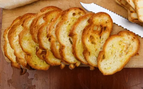 Fette dorate di pane con lievito madre, appena tostato, su un tagliere di legno. Un coltello seghettato è pronto per affettare altro pane.