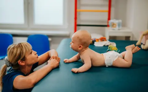 Terapeuta zachęca 3 miesięczne dziecko, które nie chce leżeć na brzuchu, do ćwiczeń na macie.