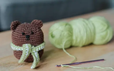 Os **sonajeros de crochet patrones gratis** son adorables. Un osito marrón con bufanda verde y blanca, junto a ovillos de lana y una aguja de crochet.