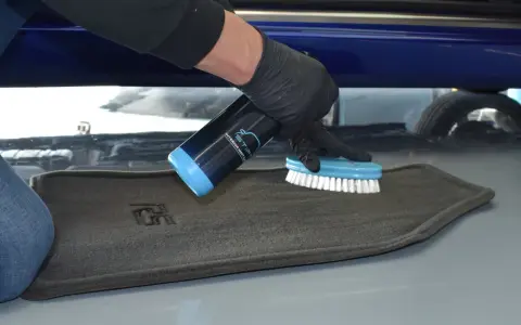 Person mit Handschuhen sprüht Reiniger auf einen grauen Autoteppich und schrubbt ihn mit einer Bürste, um ihn zu reinigen.