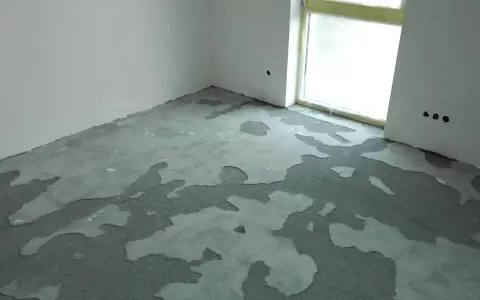 Nierównomiernie rozłożona, mokra krzywa wylewka samopoziomująca na podłodze w rogu pomieszczenia, obok okna.