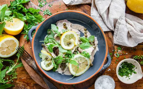 Vitello tonnato i en skål, toppad med citronskivor, kapris och ruccola. Serveras med citroner och färska örter.
