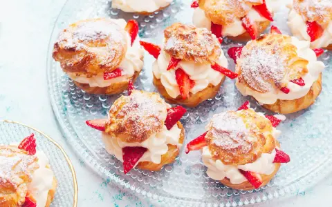 Pyszne ptysie z kremem patissiere i świeżymi truskawkami, posypane cukrem pudrem, prezentują się apetycznie na szklanej paterze.