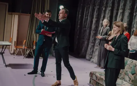 Arkadiusz Brykalski w garniturze gestykuluje na scenie, obok niego mężczyzna z teczką. W tle kobiety w eleganckich strojach.