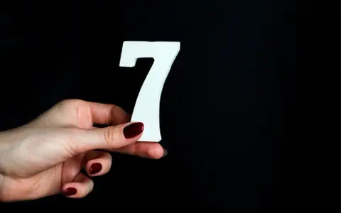 Dłoń z ciemnoczerwonym lakierem trzyma białą cyfrę 7. Może to symbolizować numerologiczną 7 mężczyzny, który jest tajemniczy i introspektywny.