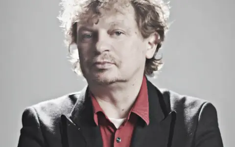 Marek Zieliński, aktor, w czarnej marynarce i czerwonej koszuli, z kręconymi włosami.