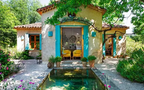 Encantadoras fachadas de casas rústicas con contraventanas turquesas y un estanque rodeado de flores.